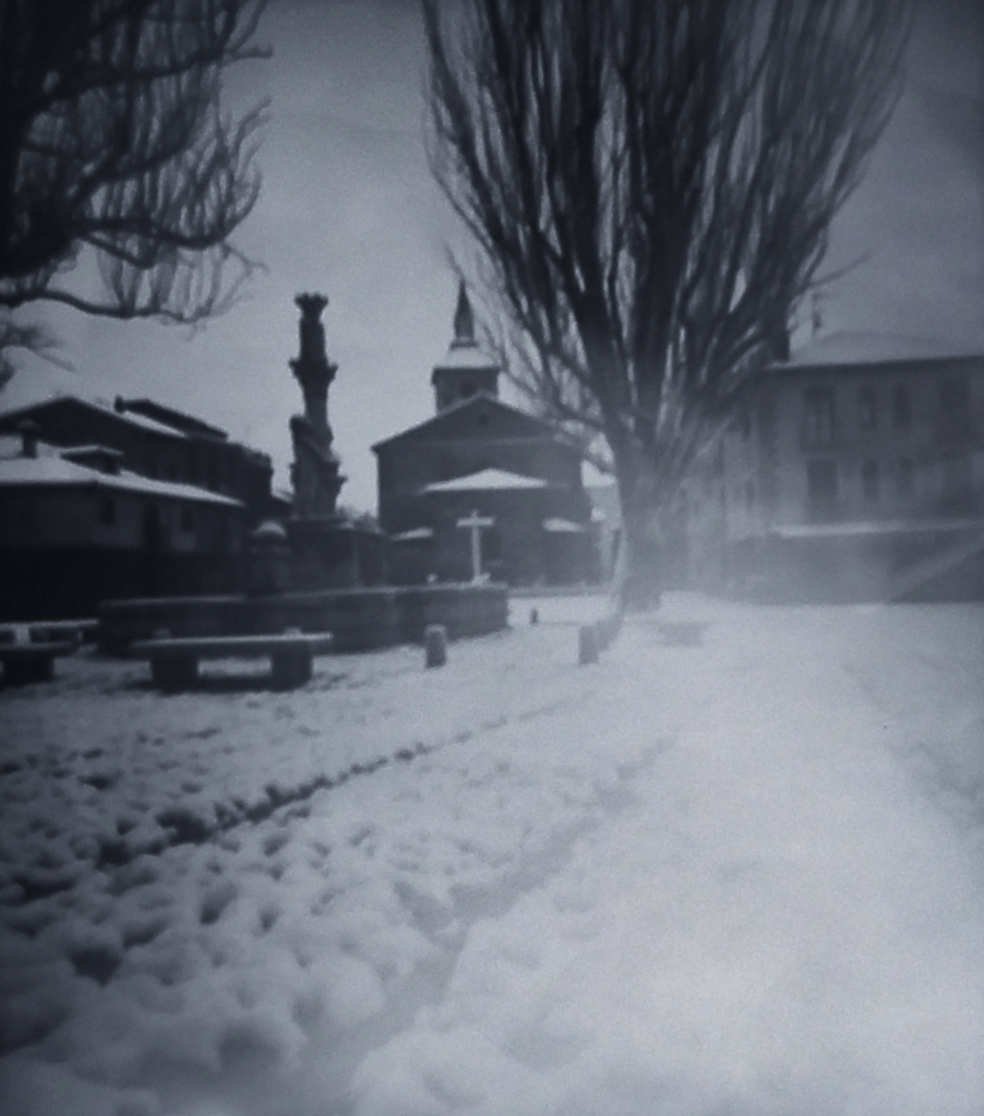 Plaza de suelo de adoquines nevada, con una iglesia al fondo, un árbol deshojado a la derecha y una fuente de piedra a la izquierda.