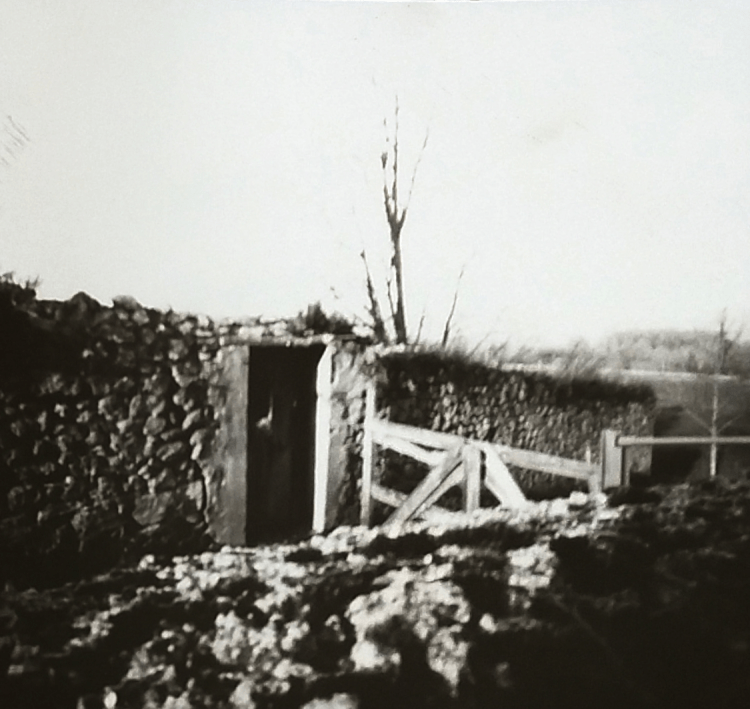 Un muro de piedra a medio derruir con una puerta de madera envejecida incrustada en él. Separando ese muro de otro, una cerca de madera impide el paso al camino existente entre ambos.